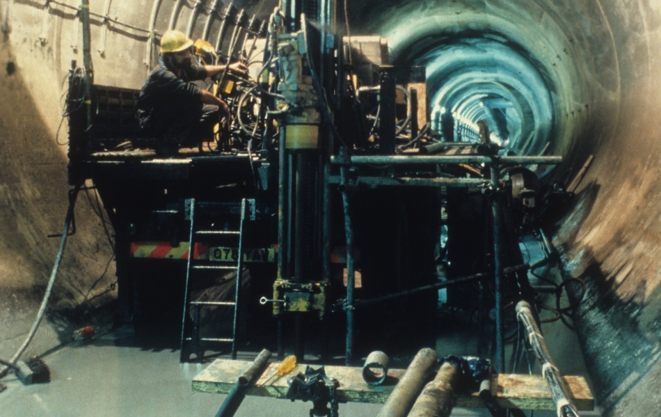 Photographie couleur montrant un chantier dans un tunnel cylindrique. Un ouvrier se trouve sur une machine, entouré de matériel et d'outils.