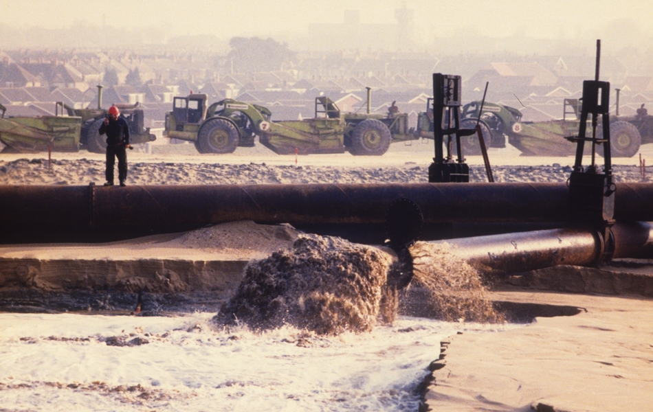 Photographie couleur montrant un chantier au bord de la mer. Au premier plan, un gros tuyau et un ouvrier ; au second plan, des engins de chantier et les toits d'une ville en arrière plan.