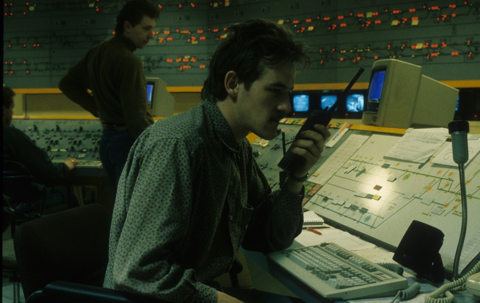 Photographie couleur montrant un poste de contrôle composé d'écrans, de voyants lumineux et de manettes. Au premier plan, un homme assis parle dans un talkie-walkie.