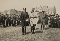 Photographie noir et blanc montrant deux hommes marchant côte à côte, l'un habillé en civil, le chapeau à la main, le second vêtu d'un uniforme militaire. Derrière eux, une foule de civils et de militaires.