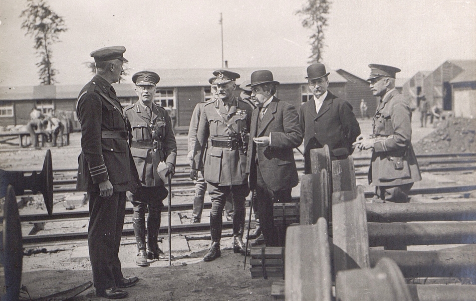 Photographie noir et blanc montrant deux civils entourés de six militaires en train de discuter autour de machines.