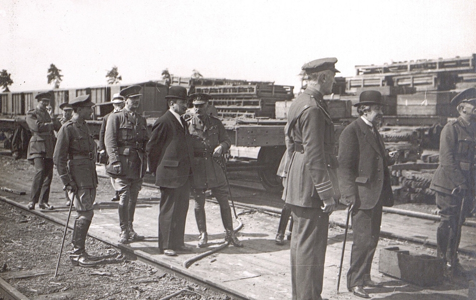 Photographie noir et blanc montrant deux civils entourés de militaires en train de discuter autour de machines et de wagons.