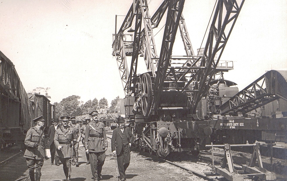 Photographie noir et blanc montrant deux civils entourés de militaires en train de discuter autour de machines et de matériel.
