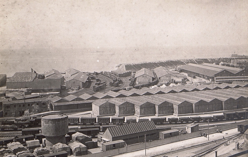 Photographie noir et blanc montrant des entrepôts et des baraquements devant un port.