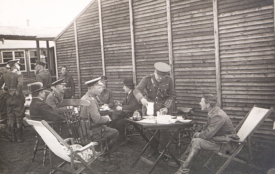 Photographie noir et blanc montrant deux civils et des militaires assis autour de tables devant des baraquements et en train de boire du thé.