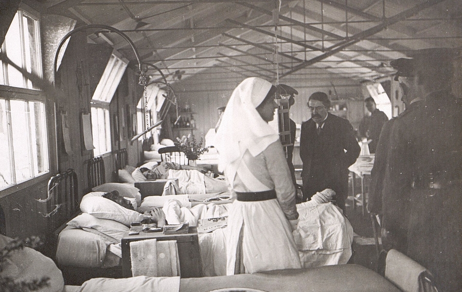 Photographie noir et blanc montrant un groupe d'hommes dans un baraquement transformé en salle d'hôpital. Au premier plan, on remarque une infirmière de dos.