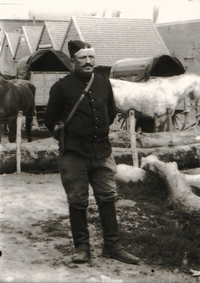 Photographie noir et blanc montrant un soldat en pied. Au second plan, on aperçoit des chevaux devant des baraquements.