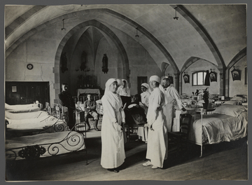 Photogrpahie noir et banc montrant l'intérieur d'un hôpital militaire installé dans une chapelle. Au premier plan posent des infirmières. Deriière elles, des lits et deux soldats.