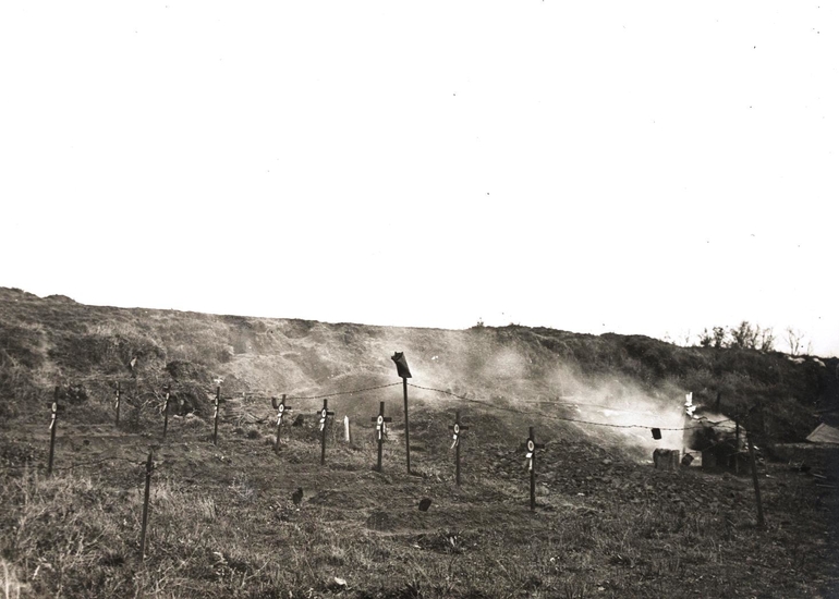 Photographie noir et blanc montrant quelques croix de bois alignées dans le sol. À l'arrière plan se dégage de la fumée.