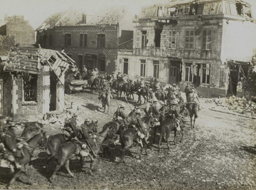 Photographie noir et blanc montrant un groupe de soldats traversant une rue de maisons détruites à cheval.