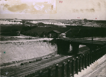 Photographie noir et blanc. Au premier plan, une voie ferrée traversée par un pont. Derrière le pont, un camp de tentes.