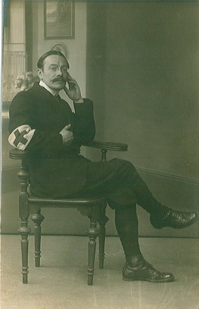Photographie noir et blanc d'un homme en costume assis sur une chaise, le coude posé sur l'accoudoir et portant un brassard orné d'une croix sur l'autre bras.