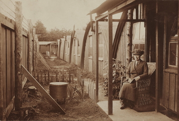 Photographie sépia montrant une femme assise devant un baraquement, un livre dans les mains.