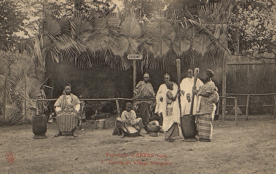 Carte postale noir et blanc d'un groupe de personnes devant une case. Deux femmes sont occupées à piler du mil.