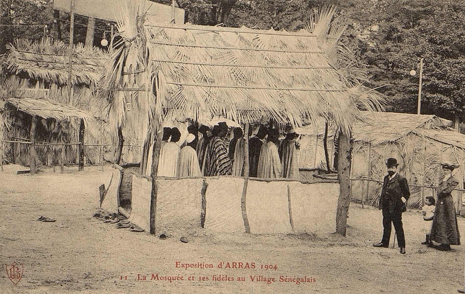 Carte postale noir et blanc d'une douzaine de personnes rassemblées dans une case ouverte.