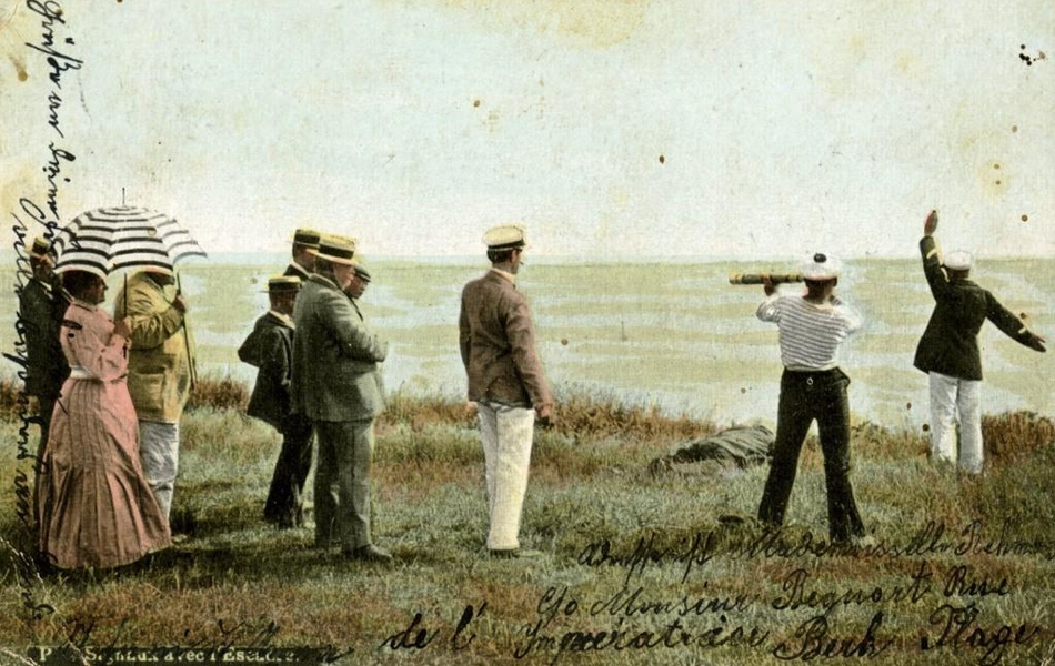 Carte postale colorisée d'un groupe de personnes au bord d'une falaise, regardant vers le large. Un homme à droite lève une main vers la mer, tandis qu'un autre derrière lui regarde dans une longue-vue