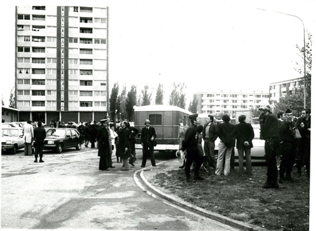 Photographie noir et blanc d'une rue de cité. Une foule de badauds et d'agents se presse au pied d'un immeuble.