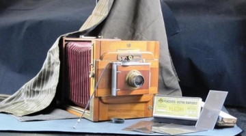 Photographie couleur montrant une boîte photographique carrée, en bois et en cuir (pour la partie en accordéon), sur laquelle repose un drap de photographe. À droite de l'appareil est posée une boîte sur laquelle on lit "plaques ultra-rapide". Son contenu est présenté au premier plan, il s'agit de plaques de verre.