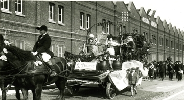 Photographie noir et blanc d'un char tiré par un cheval et portant la pancarte "La Brasserie", passant devant une usine. Des jeunes filles habillées en moissonneuses s'y tiennent debout, entourées de branches de houblon et de fûts. Derrière suit un cortège.