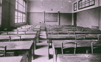 Photographie noir et blanc d'une classe vide meublée de pupitres, de chaises et d'un tableau noir.