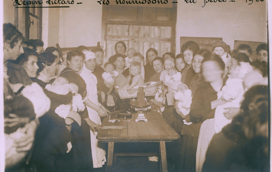 Photographie noir et blanc montrant des femmes et des bébés entourant une table sur laquelle se trouve une balance.