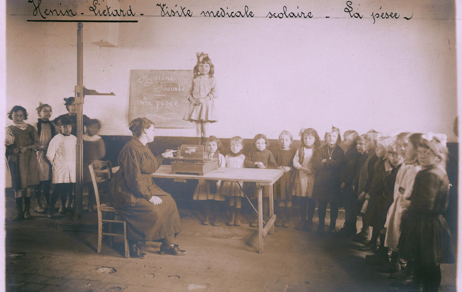 Photographie noir et blanc d'élèves en file indienne dans une classe d'école. Ils passent à tour de rôle devant le bureau de l'institutrice qui les pèse et mesure à tour de rôle.