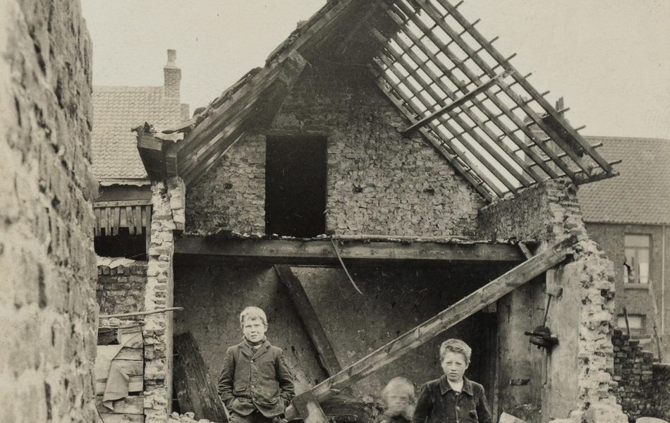 Photographie noir et blanc montrant trois enfants debout parmi des gravas de maisons détruites.