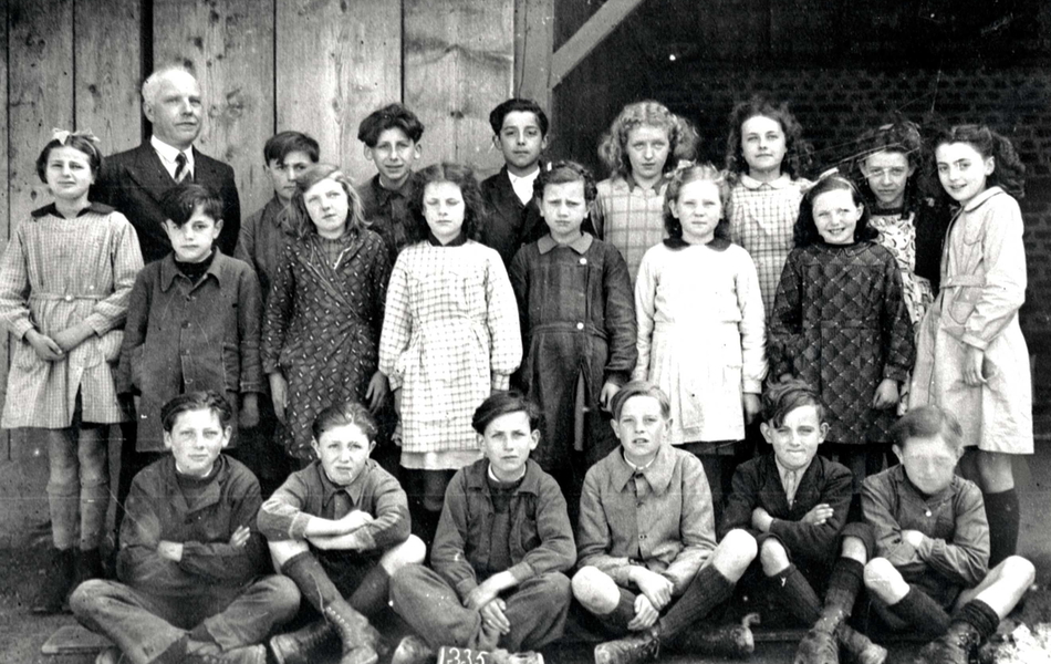 Photographie de classe noir et blanc montrant de jeunes garçons et de jeunes filles posant sur trois rangs aux côtés de leur professeur.