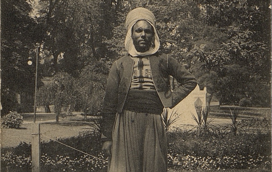 Carte postale noir et blanc d'un homme en tenue de zouave, une main sur la hanche, posant devant des pelouses fleuries.