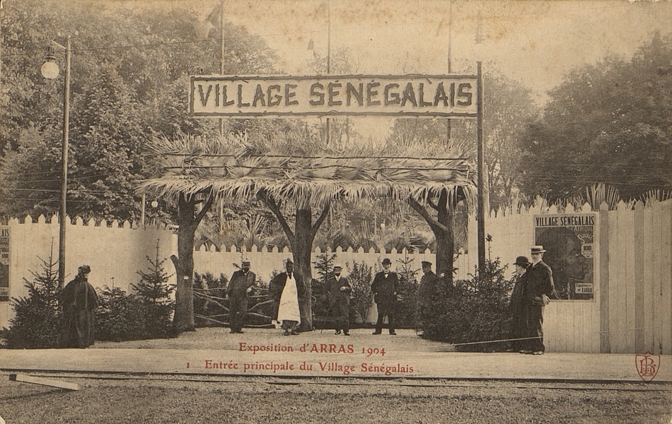 Carte postale noir et blanc montrant un porte en branches et palmes, au-dessus de laquelle se trouve un panneau où il est écrit "village sénégalais". Devant, des européens en pardessus et des africains en habit traditionnel posent.