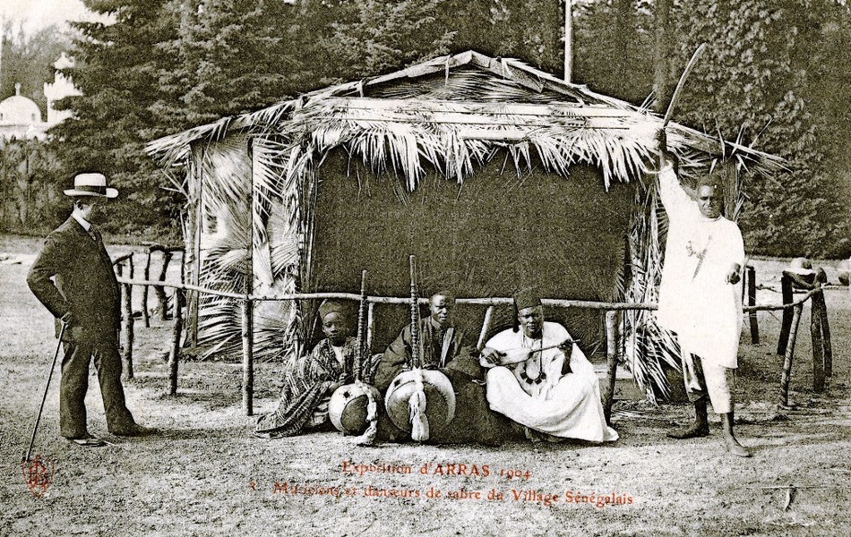 Carte postale noir et blanc d'un groupe d'hommes posant devant une case faite de feuilles de palmiers. À gauche se trouve un homme vêtu d'un complet et tenant une canne. Puis viennent trois hommes noirs accroupis et tenant des instruments de musique, et enfin un homme de couleur, debout et brandissant un sabre au-dessus de sa tête.