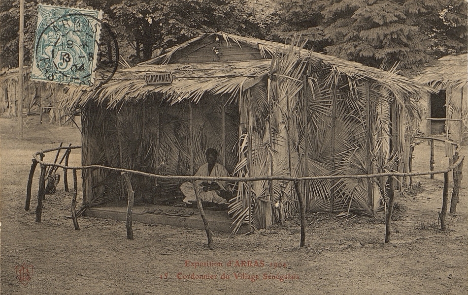 Carte postale noir et blanc d'un homme assis dans une case ouverte. Au-dessus est accroché le panneau "cordonnier".