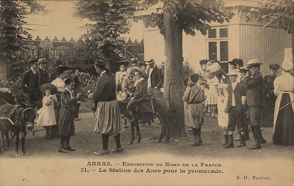 Carte postale noir et blanc où l'on voit un groupe de personnes debout entre les arbres. Certains regardent des ânes tirés par des guides et montés d'enfants.