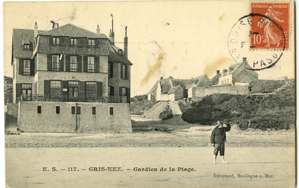 Carte postale en noir et blanc de la plage bordée de quelques maisons et de végétation. Au milieu se tient un homme, pieds nus et le pantalon relevé, une main se protégeant du soleil et regardant au loin