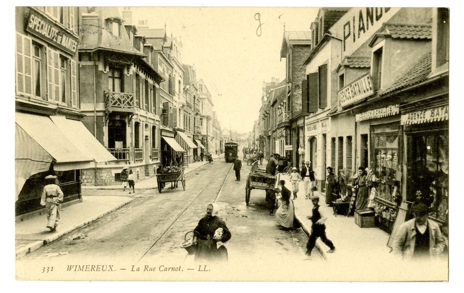 Carte postale en noir et blanc de la rue Carnot, avec le tramway au milieu. Quelques carrioles sont garées le long de cette rue commerçante très fréquentée