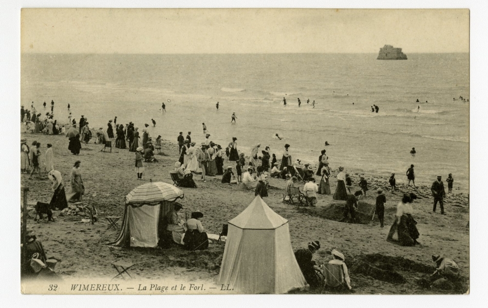 Carte postale en noir et blanc de la plage en pleine saison. On y voit quelques tentes de plage, des enfants creusant dans le sable, des promeneurs le long de l’eau et des baigneurs. Au large, on distingue le fort