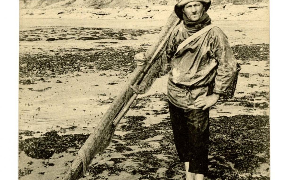 Carte postale en noir et blanc d’un homme, en pied, posant sur la plage à marée basse. Pieds nus, il porte une vareuse, un chapeau de pêcheur, un panier en osier en bandoulière et s’appuie contre un mât