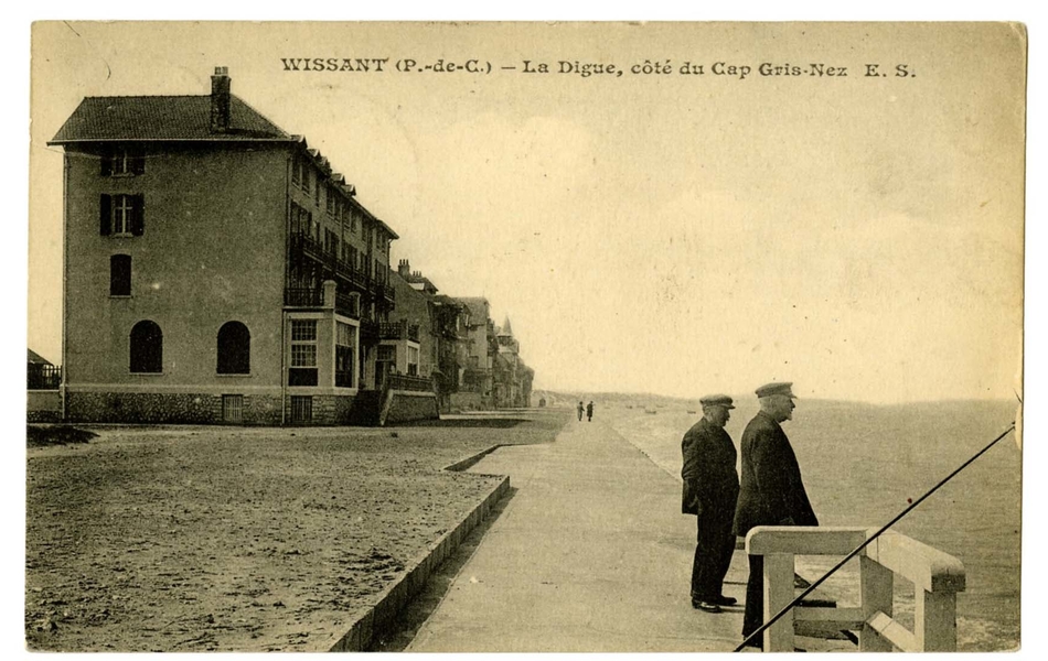 Carte postale en noir et blanc de la digue occupée par quelques promeneurs contemplant le large. Quelques maisons bordent la jetée