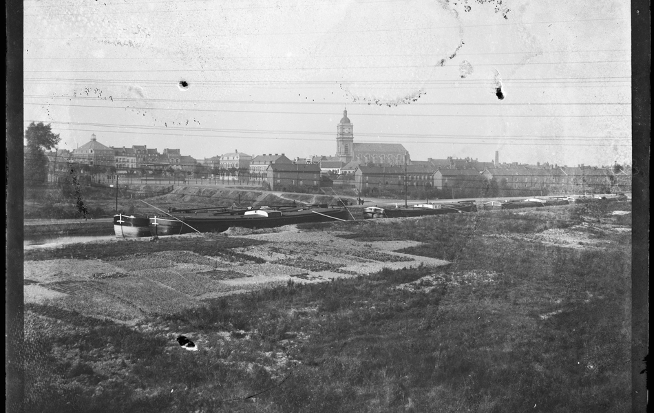 Photographie noir et blanc montrant les rives d'un canal sur lequel sont amarrées des péniches. À l'arrière plan se dessine une ville.