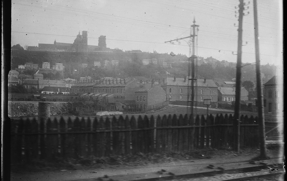 Photographie noir et blanc montrant un groupe de bâtiments sur une colline, devant une voie ferrée.