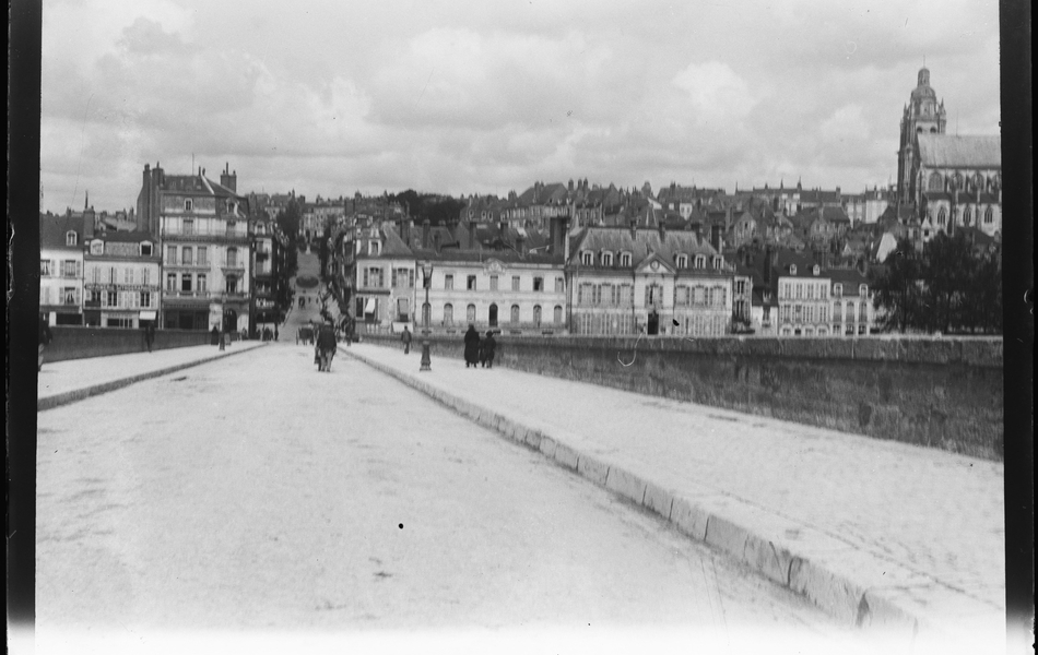 Photographie noir et blanc montrant une route pavée sur un pont. À l'arrière plan se dresse la ville.