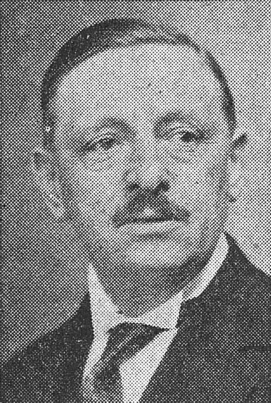 Portrait noir et blanc montrant un homme portant une petite moustache et une cravatte sous un costume sombre.