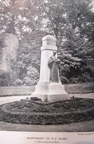 Photographie noir et blanc d'une colonne sur laquelle repose le buste d'Ernest Hamy. Devant cette colonne on voit une statue de femme tenant un crâne dans sa main. Le monument se trouve dans un parc, au milieu d'un parterre de fleurs.
