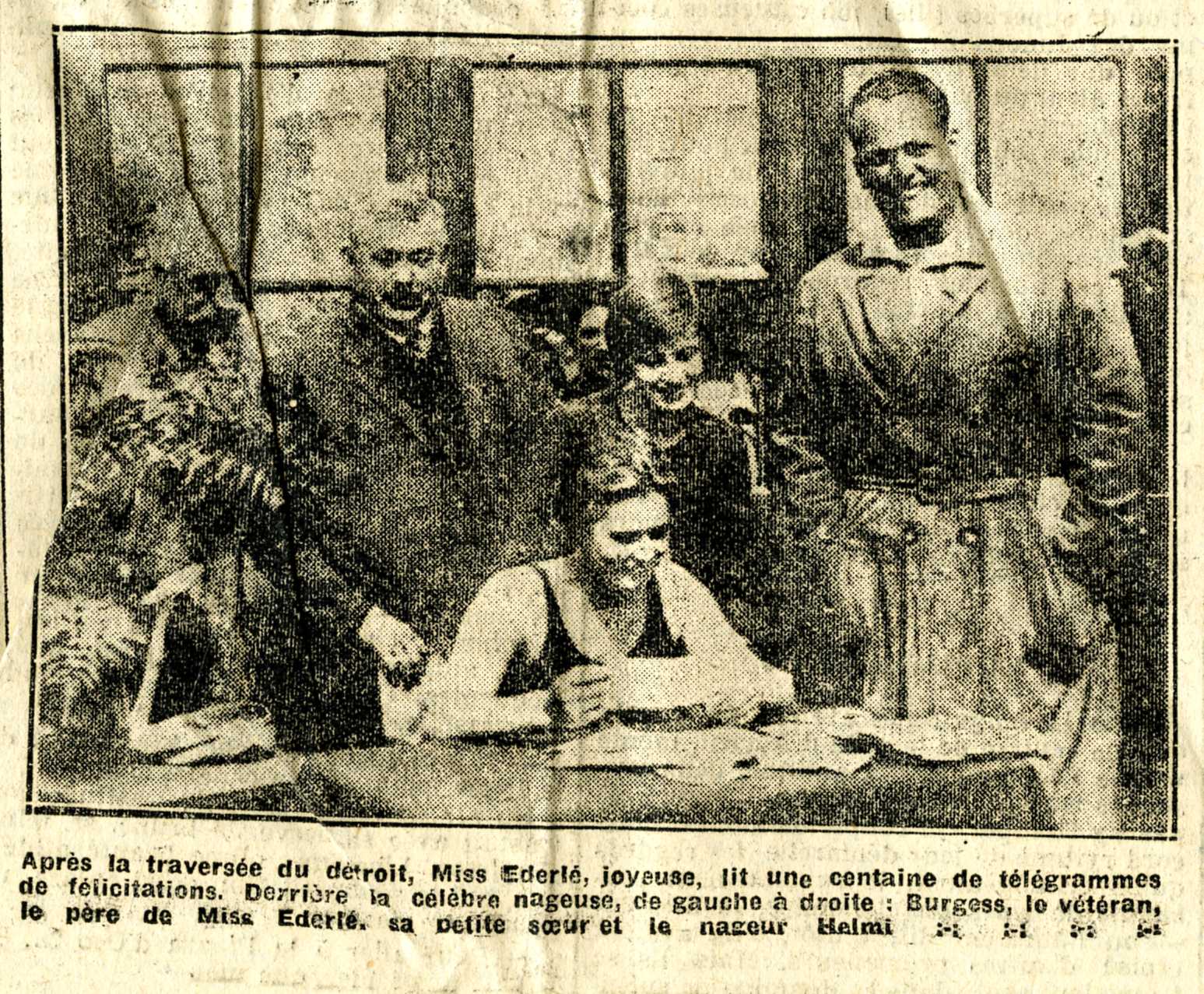 Photographie noir et blanc d'une femme en maillot de bain assise en train de lire, entourée de quatre personnes debout derrière elle. Légende : Après la traversée du détroit, Miss Éderlé, joyeuse, lit une centaine de télégrammes de félicitations. Derrière la célèbre nageuse, de gauche à droite : Burgess, le vétéran, le père de Miss Éderlé, sa petite soeur et le nageur Helmi.