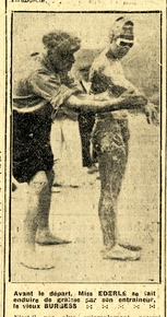 Photographie noir et blanc d'une femme en maillot de bain portant lunettes de plongée et bonnet de bain qui regarde un homme qui lui passe la main sur le bras. Légende : Avant le départ, Miss Éderlé se fait enduire de graisse par son entraîneur, le vieux Burgess.