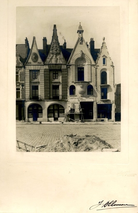 Photographie noir et blanc montrant les façades de quelques maisons situées sur une place pavée en travaux. Devant une maison est dressé un échafaudage sur lesquel deux ouvriers s'activent. Les façades sont ornées de bas-reliefs et frontons en forme de visages, soleils et bouquets.