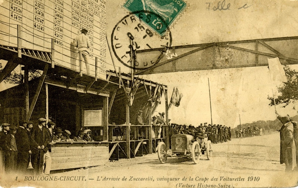 Carte postale noir et blanc d’une ligne d’arrivée de course automobile. Au premier plan, on voit une tribune de juges devant laquelle stationne une voiture portant le numéro deux. Au-dessus, un homme inscrit les résultats sur un grand panneau.