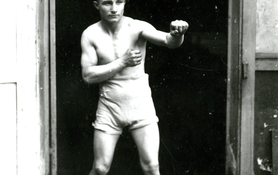 Photographie noir et blanc d’un boxeur en pied dans l’embrasure d’une porte, torse nu, vêtu d’un short et de chaussures, les poings levés.