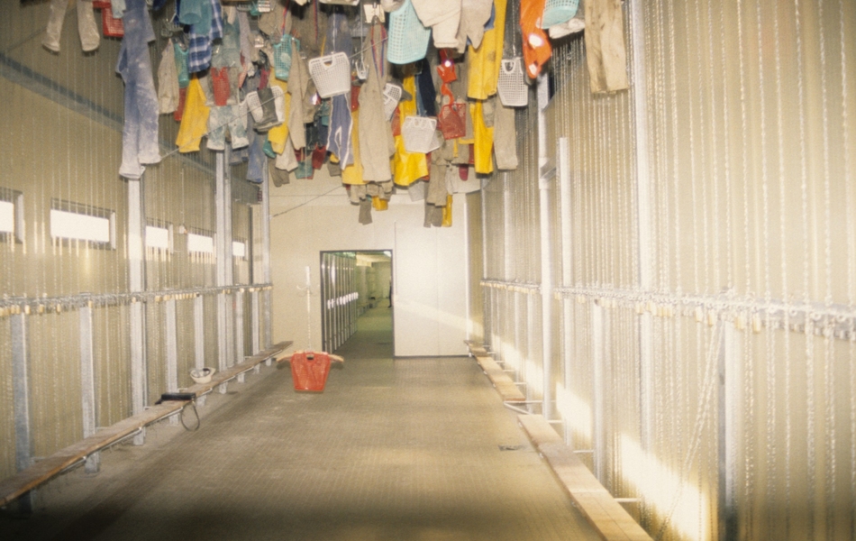 Photographie couleur montrant une salle vide, avec juste un banc. Au plafond, on voit des paniers et du linge suspendus.