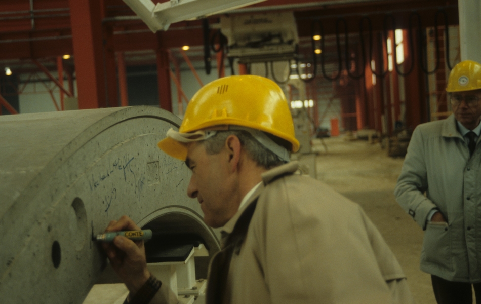 Photographie couleur montrant un homme portant un casque de chantier jaune en train d'écrire sur une structure en béton, dans un entrepôt d'usine.